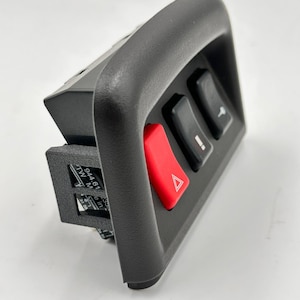 Könnte beinhalten: Eine schwarze Kunststoff-Bedienblende mit drei Schaltern und einem roten Warnblinklichtknopf. Die Schalter sind schwarz mit weißen Symbolen. Die Blende hat einen gebogenen Griff und eine rechteckige Basis.