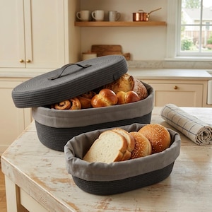 May include: Two oval bread baskets, one with a lid, filled with various types of bread. The baskets are dark grey with a grey fabric lining. The lid has a handle. The baskets sit on a light-coloured wooden table.