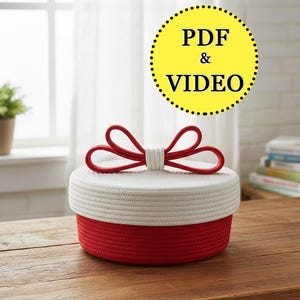 May include: A two-tone storage basket with a red base and a white lid, topped with a red bow. The text "PDF & VIDEO" is in a yellow circle. The basket is on a wooden surface. The basket's design suggests it is handmade.