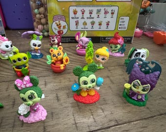 May include: A collection of colorful character figurines, including Mickey and Minnie Mouse, and other Disney-themed figures. The figurines are set on a wooden surface, with a box in the background.
