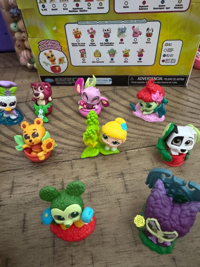 May include: A collection of colorful Fantasy Florals figurines featuring Disney characters. The figurines include Winnie the Pooh, Stitch, and others, each with floral elements. The packaging is visible in the background.