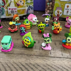May include: A collection of colorful miniature figurines, including characters like an alien, a bear in a flower pot, and a bus. The figurines are set on a wooden surface, with some packaging visible in the background.