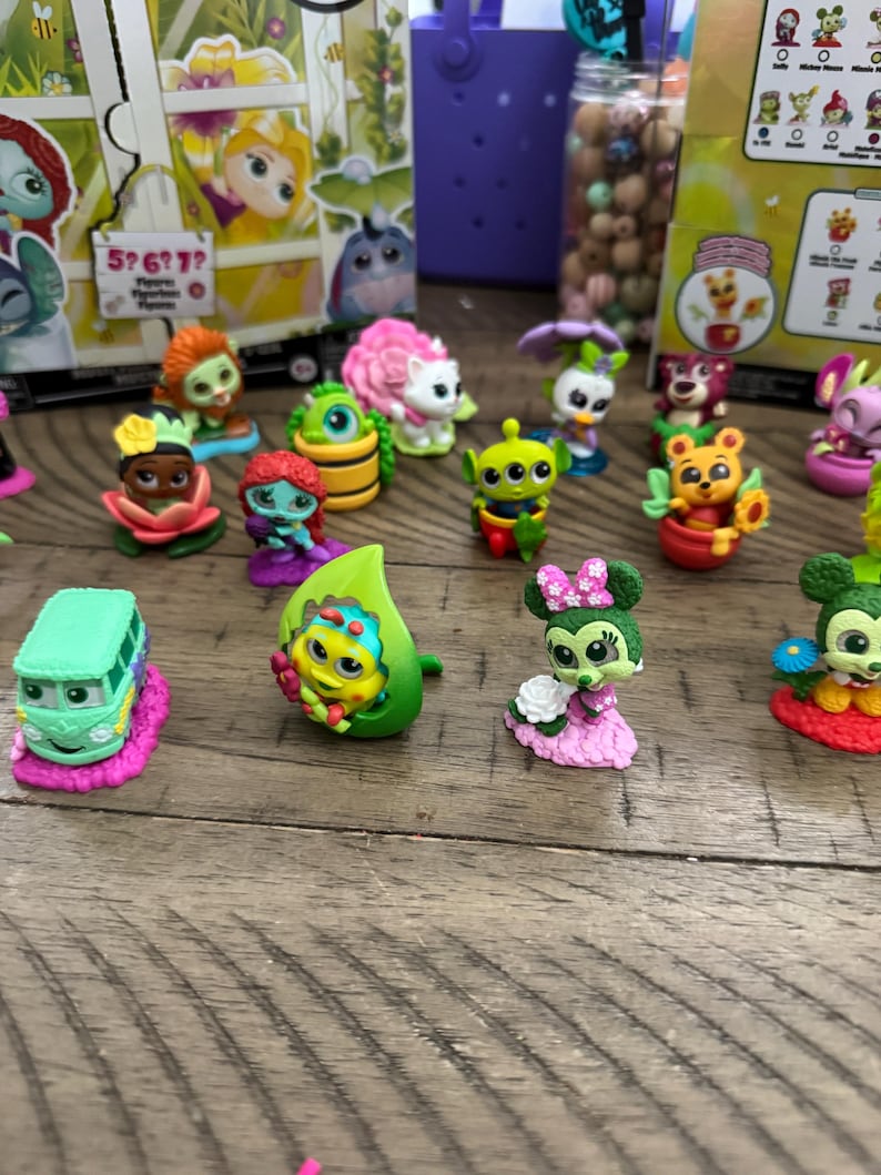May include: A collection of colorful miniature figurines, including characters like an alien, a bear in a flower pot, and a bus. The figurines are set on a wooden surface, with some packaging visible in the background.