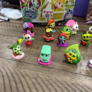 May include: A collection of small, colorful character figurines, including a green van, a pink bunny, and various other figures. The figurines are set on a wooden surface, with a box featuring cartoon characters in the background.
