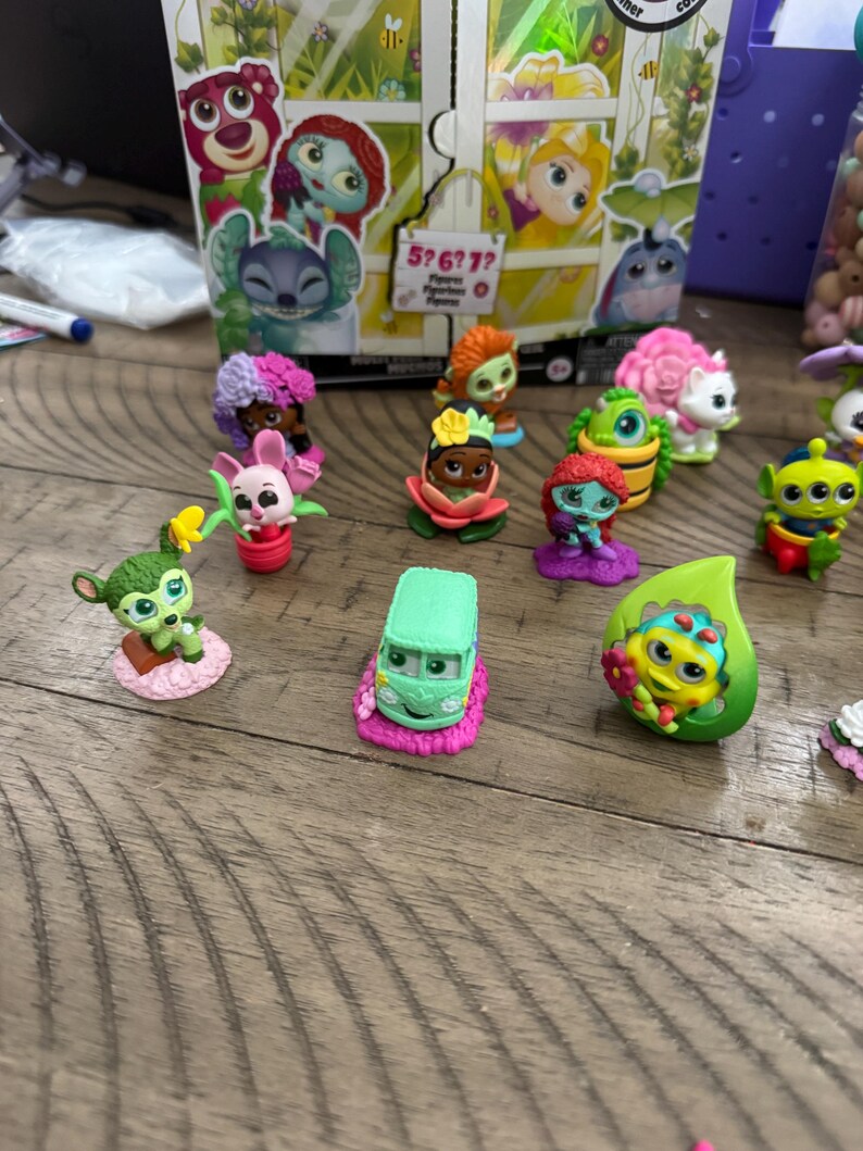 May include: A collection of small, colorful character figurines, including a green van, a pink bunny, and various other figures. The figurines are set on a wooden surface, with a box featuring cartoon characters in the background.