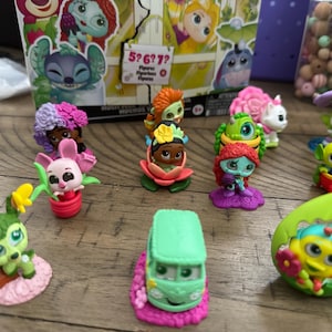 May include: A collection of colorful character figurines, including a green alien, a pink bunny, and a green van. The figurines are set on a wooden surface, with a box featuring cartoon characters in the background.