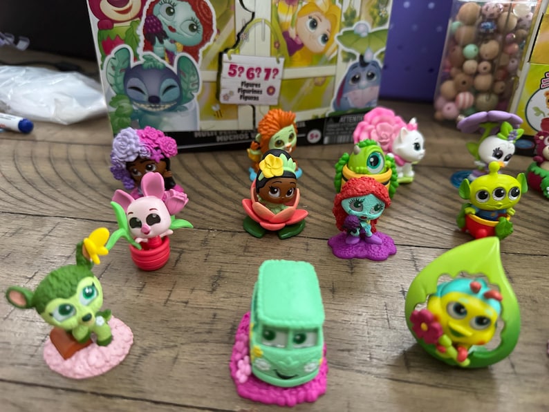 May include: A collection of colorful character figurines, including a green alien, a pink bunny, and a green van. The figurines are set on a wooden surface, with a box featuring cartoon characters in the background.