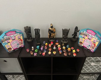 May include: A collection of small, colorful, plastic figurines of Disney characters. The figurines are arranged in a row on a dark brown shelf. There are two boxes of Disney Doorables on the shelf, one on each end. The boxes are blue and pink with the words "Disney Doorables" printed on them. There are also two black and gold tiki statues on the shelf.