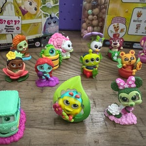 May include: A collection of colorful character figurines, including a green alien, a bear in a pot, and a Minnie Mouse figure with a pink bow. The figurines are on a wooden surface, with packaging visible in the background.