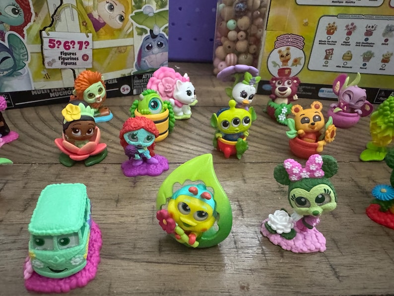 May include: A collection of colorful character figurines, including a green alien, a bear in a pot, and a Minnie Mouse figure with a pink bow. The figurines are on a wooden surface, with packaging visible in the background.