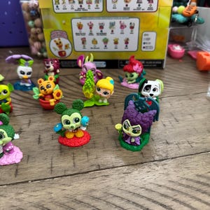 May include: A collection of colorful miniature figurines, including characters with floral and animal themes, are displayed on a wooden surface. A box with character illustrations is in the background. The figurines have bright colors and detailed designs.