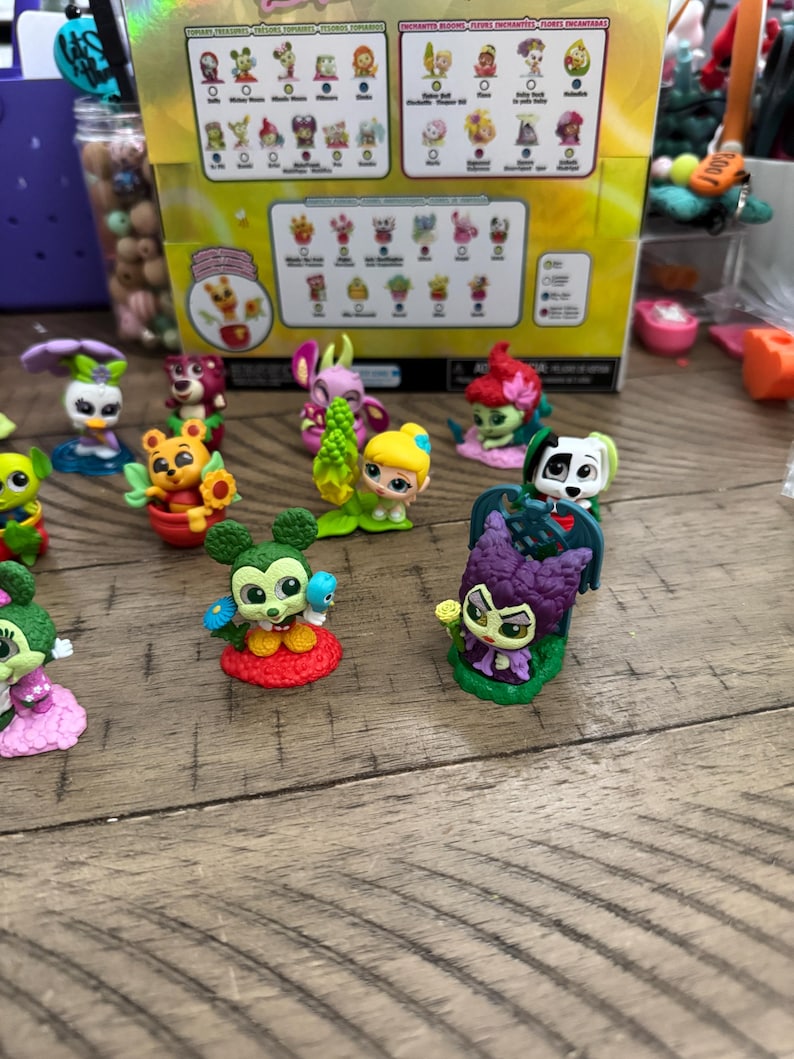 May include: A collection of colorful miniature figurines, including characters with floral and animal themes, are displayed on a wooden surface. A box with character illustrations is in the background. The figurines have bright colors and detailed designs.