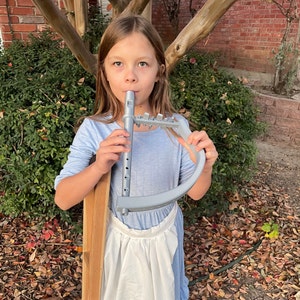 May include: A young person playing a gray plastic musical instrument, resembling a flute, with a curved handle. The instrument is held in front of a light blue dress and a white apron. The background includes green foliage and a brick wall.