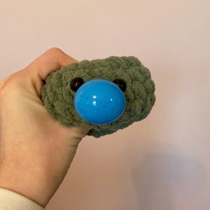 May include: A handmade, green crocheted plush toy with a large, blue, spherical nose and two small, black eyes. The toy is held in a hand against a light pink background. The plush has a textured, bumpy surface.