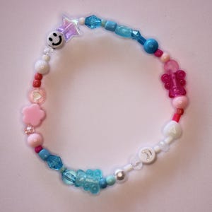 May include: A beaded bracelet featuring a variety of colourful shapes including a white smiley face, a translucent purple star, a pink flower, a pink butterfly, a blue bear, and a white bead with a silver moon symbol, interspersed with round and faceted beads.
