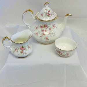 May include: A white porcelain tea set featuring a pink rose floral design and gold edging. The set comprises a teapot, a creamer jug, and a teacup. The teapot has a lid and a curved handle. The set is displayed on a white surface.
