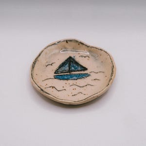 Peut inclure: Un petit plat en céramique rond et blanc cassé avec un bord irrégulier formé à la main. Le centre présente une illustration peinte à la main d'un voilier avec une coque et une voile bleues, entouré de dessins au trait noir de vagues et d'oiseaux.