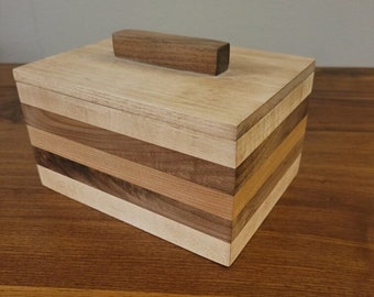 May include: A wooden box with a layered design, featuring different wood types. The box has a lid with a rectangular wooden handle.