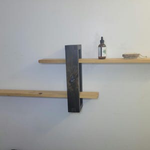 May include: A wooden shelf unit with two shelves and a black metal support. The shelves are made of natural wood and the support is painted black. There is a small bottle and a brush on the top shelf.