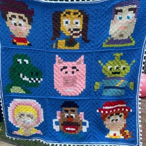 以下が含まれることがあります： 手作りの青いかぎ針編みの毛布で、アニメ映画トイ・ストーリーのキャラクターのピクセル化された画像が特徴です。 毛布には白い縁取りがあり、ウッディ、バズ・ライトイヤーなどのキャラクターが含まれています。 屋外に展示されています。