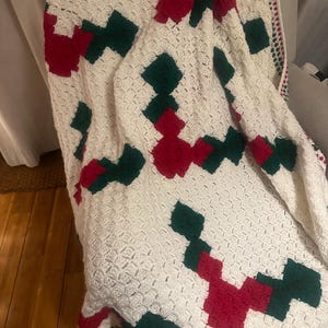 May include: A white crocheted blanket with a green and red geometric pattern. The blanket is folded and draped over a chair.