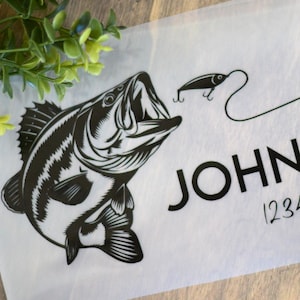 May include: A black and white graphic design on a white background. The design features a fish, fishing rod, and the name "JOHNSON" with the address "1234 Main Street". The image is on a wooden surface with green plants.