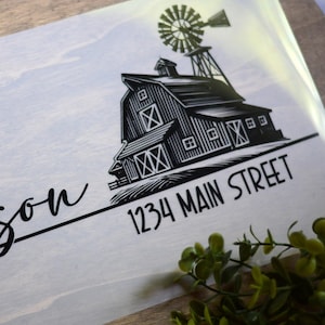May include: A wooden sign with the name "Johnson" in elegant script. A black and white illustration of a barn with a windmill is featured, along with the address "1234 Main Street". Greenery accents the sign's edges.