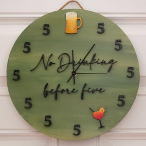 May include: Round, green wooden clock with the words "No Drinking before five" in black script. The clock has black numbers and decorative images of a beer mug and a cocktail. A rope is attached for hanging.
