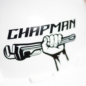 Puede incluir: Un casco blanco con un logotipo en blanco y negro. El logotipo muestra una mano sosteniendo una llave inglesa y la palabra "CHAPMAN" en letras negras y negritas.