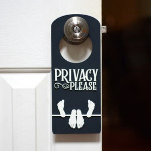 May include: A black door hanger with the text "Privacy Please" in white lettering. The hanger has a white silhouette of two feet sticking out from the bottom.