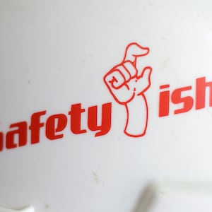 Puede incluir: Un casco blanco con un logotipo rojo que dice "safety ish" y una mano de dibujos animados roja con un dedo apuntando hacia arriba.