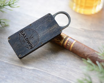 Puede incluir: Un cortapuros de madera con un abridor de anillos y un logotipo en blanco y negro que dice "Baldwin 1927". El cortapuros está sobre una superficie de madera junto a un puro marrón.