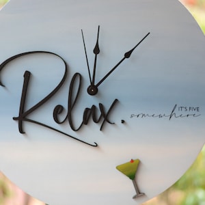 May include: A round, white clock with a blue ombre effect. The clock face says "Relax. It's five somewhere" and features a black cocktail glass with a green drink and a red cherry. The clock hands are black.