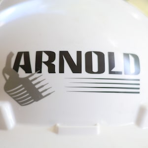 Puede incluir: Casco blanco con la palabra "ARNOLD" impresa en letras negras. Una silueta gris de una excavadora de construcción está a la izquierda del texto.