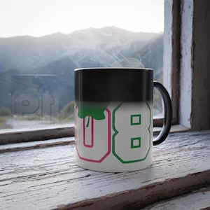 Puede incluir: Una taza que cambia de color, negra y blanca, con los números "08" en fuente blanca delineada en rosa y verde. Sale vapor de la taza, que está sobre un alféizar de madera desgastada con vistas a la montaña al fondo.