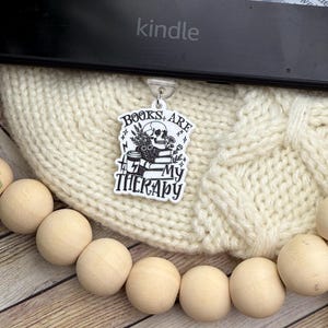 May include: A black and white charm with the text "BOOKS ARE MY THERAPY" and a skull design, hanging from a Kindle e-reader. The charm is set against a cream-colored knitted background, with wooden beads in the foreground.