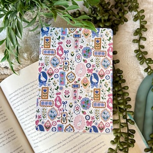 May include: A patterned notebook featuring a repeating design of framed floral and bird illustrations in pink, blue, and peach. The notebook is open on a book with text.