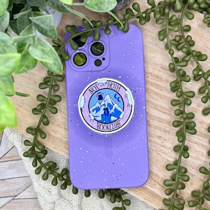 May include: A purple speckled phone case with a pop-up grip featuring the text "NOT HOTH BOOKCLUB" and a mountain scene. The phone case is surrounded by green plants and rests on a wooden surface.