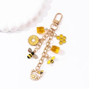 May include: A gold-toned metal keychain featuring multiple charms including two yellow honey pots, a yellow daisy, a honeycomb shape, two bees with black and yellow stripes, and a heart-shaped charm decorated with a small bee and white floral accents.