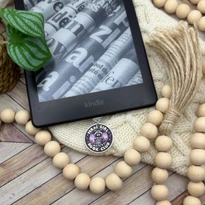 May include: A black and white book club charm with a raccoon wearing glasses and holding a book. The charm is attached to a wooden bead necklace. The charm has the text "Feral Girl Book Club" on it.