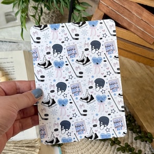 May include: A rectangular notebook with a repeating pattern of hockey skates, helmets, hockey sticks, and text that reads "I like my books spicy & coffee icy." The design includes pink bows, snowflakes, and stars.