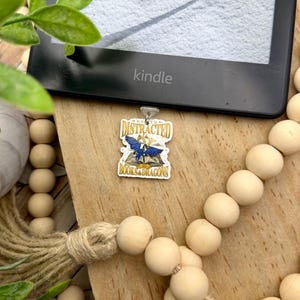 May include: A blue and gold dragon charm with the text "Easily Distracted by Books & Dragons". The charm is attached to a silver chain and is hanging from a Kindle e-reader.