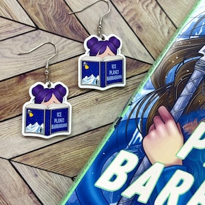 May include: A pair of earrings featuring a book with the title "Ice Planet Barbarians" and a cartoon girl reading it. The earrings are silver and have a white background with blue and green accents.