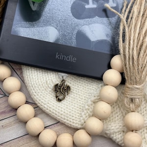 May include: A black Kindle e-reader with the word "kindle" printed on the bottom. A cream-colored knitted cover with a bronze dragon charm is attached. A wooden bead garland and a bundle of twine are also visible.