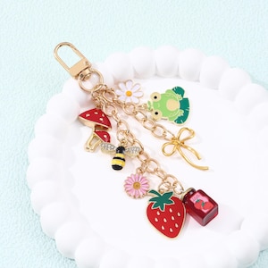 May include: A gold-colored keychain with multiple charms. Charms include a red mushroom, a bee, a pink flower, a daisy, a green frog, a gold bow, a red strawberry, and a jar of jam. The charms are attached to a gold chain.