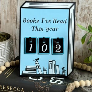 May include: A light blue book-shaped counter with the text "Books I've Read This year" and the number "102". The counter has a black and white border and a decorative illustration of books and a bird.