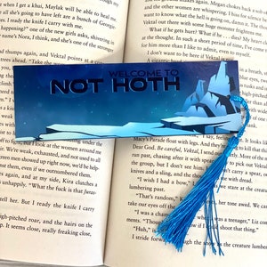 May include: A blue bookmark with a tassel, featuring the words "Welcome to Not Hoth" in white text on a dark blue background. The bookmark is partially visible on a page of a book with text about a fictional world.