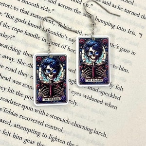 May include: A pair of rectangular earrings featuring a skeleton with blue hair and glasses reading a book. The earrings have a floral border and the text "THE READER". The earrings hang from silver hooks.