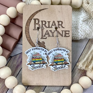May include: A pair of dangle earrings with a book theme. Each earring features a colorful stack of books with the text "Easily Distracted By Books". The earrings are on a wooden display with the text "Briar Layne".