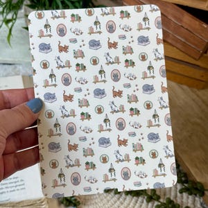 May include: A rectangular card with a repeating pattern of cats, books, and potted plants in soft colors. The cats are depicted in various poses, including sleeping and playing. The overall design has a whimsical, vintage aesthetic.